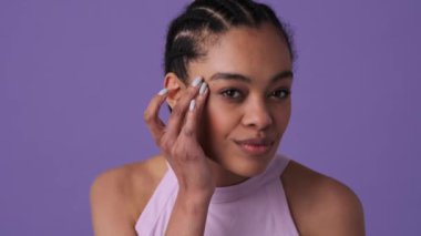 Memnun genç bir kadın kameraya bakıyor. Stüdyonun menekşe arka planında yalnız duran aynaya bakıyor.