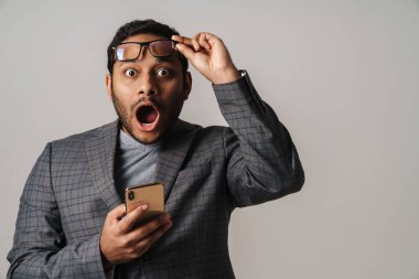 Asyalı şaşkın işadamı kameraya bağırıyor ve cep telefonunu gri arka planda tutuyor.