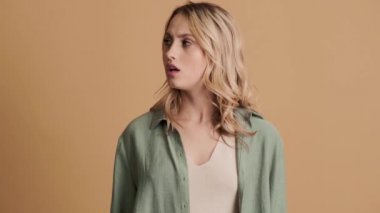 Kızgın genç bir kadın, stüdyoda bej arka planda negatif bir şekilde dururken ellerini çapraz yapıyor.