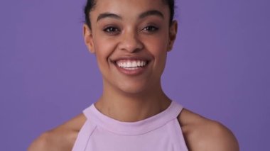 Gündelik giysiler giyen çekici bir Afrikalı genç kadın, stüdyodaki menekşe arka planında tek başına gülümsüyor.