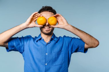 Mavi arka planda izole edilmiş mandalinalarla eğlenirken gülümseyen yakışıklı adam.