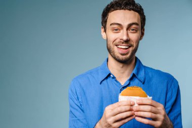 Mavi arka planda saklanmış hamburgerle poz verirken gülümseyen neşeli yakışıklı adam.