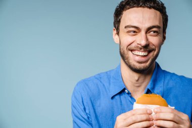 Mavi arka planda izole edilmiş hamburgerle poz verirken gülen neşeli yakışıklı adam.