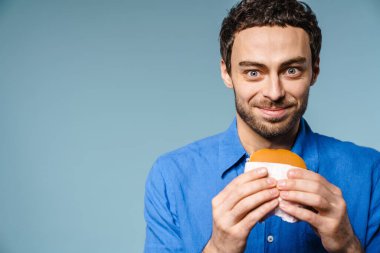 Mavi arka planda izole edilmiş hamburgerle poz verirken gülümseyen neşeli yakışıklı adam.