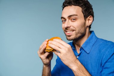 Mavi arka planda izole edilmiş hamburger yerken gülümseyen yakışıklı adam.