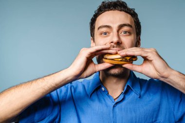Heyecanlı yakışıklı, aç adam mavi arka planda kamerada hamburger yiyor.