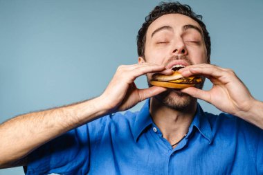 Heyecanlı yakışıklı, aç adam mavi arka planda kamerada hamburger yiyor.