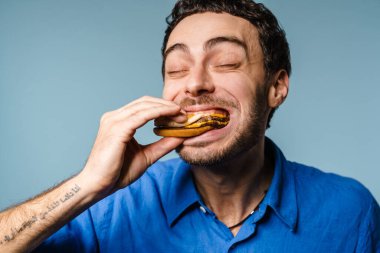 Heyecanlı yakışıklı, aç adam mavi arka planda kamerada hamburger yiyor.