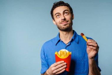 Mavi arka planda izole edilmiş ketçaplı patates kızartması yerken gülümseyen yakışıklı adam.