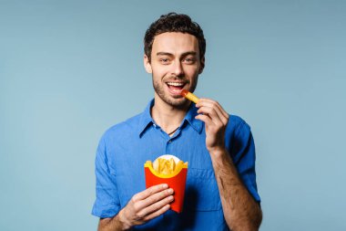 Mavi arka planda izole edilmiş ketçaplı patates kızartması yerken gülümseyen yakışıklı adam.