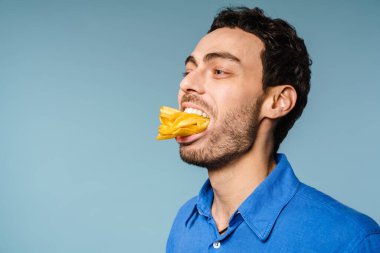 Ağzında patates kızartmasıyla poz veren mutlu yakışıklı adam mavi arka planda izole edilmiş.