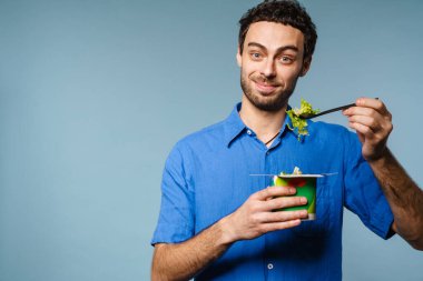 Mavi arka planda salata yerken gülümseyen yakışıklı adam.