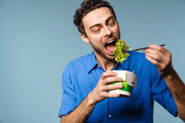 Heyecanlı yakışıklı aç adam mavi arka planda izole edilmiş salata siparişi yiyor.
