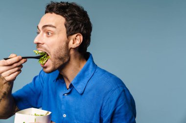 Heyecanlı yakışıklı aç adam mavi arka planda izole edilmiş salata siparişi yiyor.