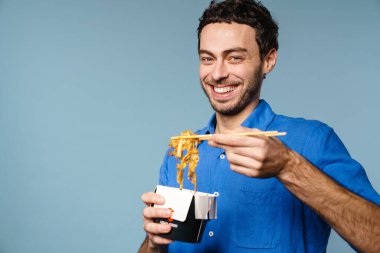 Asyalı erişteleri yerken gülümseyen neşeli yakışıklı adam mavi arka planda izole edilmiş.