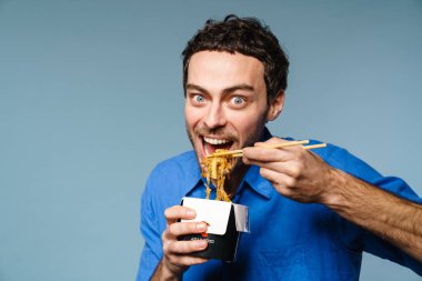 Asyalı erişte yiyen heyecanlı yakışıklı adam mavi arka planda izole edilmiş.