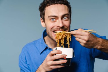 Asyalı erişte yiyen heyecanlı yakışıklı adam mavi arka planda izole edilmiş.