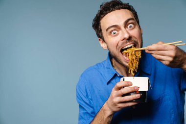 Asyalı erişte yiyen heyecanlı yakışıklı adam mavi arka planda izole edilmiş.