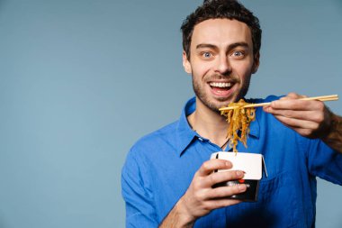 Asyalı erişteleri yerken gülümseyen neşeli yakışıklı adam mavi arka planda izole edilmiş.