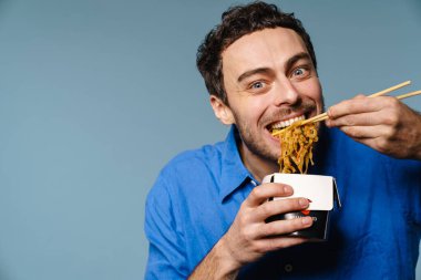 Asyalı erişteleri yerken gülümseyen neşeli yakışıklı adam mavi arka planda izole edilmiş.