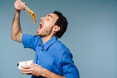 Asyalı erişte yiyen heyecanlı yakışıklı adam mavi arka planda izole edilmiş.