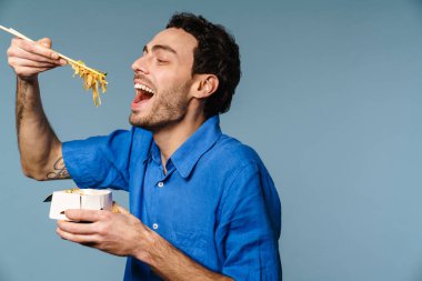 Asyalı erişte yiyen heyecanlı yakışıklı adam mavi arka planda izole edilmiş.