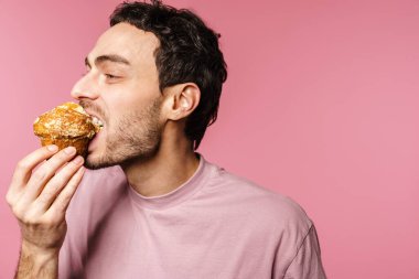 Mutlu yakışıklı adam pembe arka planda gözleri kapalı kek yiyor.