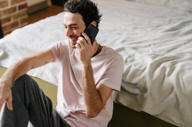 Yakışıklı, traşsız, neşeli adam. Yatak odasında yerde otururken cep telefonuyla konuşuyor.