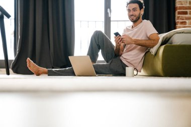 Yatak odasında yerde otururken cep telefonu ve dizüstü bilgisayar kullanan yakışıklı adam.