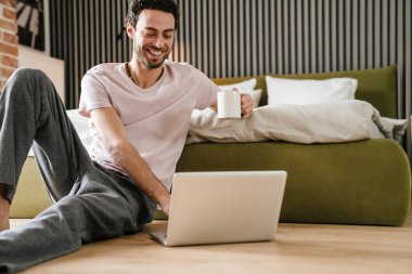 Yakışıklı gülen adam yatak odasında yerde otururken dizüstü bilgisayar kullanıyor ve kahve içiyor.