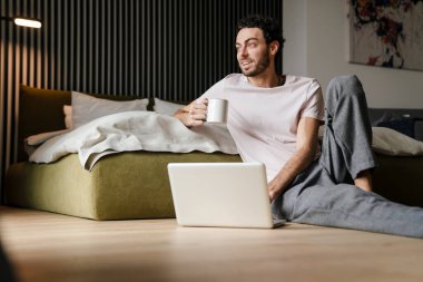 Yakışıklı adam yatak odasında yerde otururken dizüstü bilgisayar kullanıyor ve kahve içiyor.