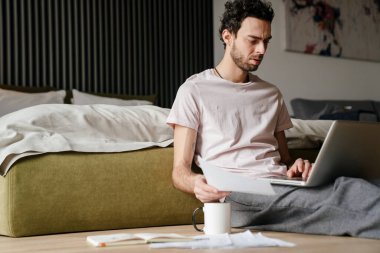 Yatak odasında yerde otururken bilgisayarla ve kağıtlarla çalışan kafası karışık yakışıklı bir adam.