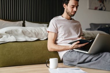 Yatak odasında dizüstü bilgisayarla çalışırken notlar yazan yakışıklı adam.