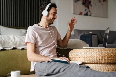 Kulaklıklı gülümseyen adam, yatak odasındaki laptopta video görüşmesi yaparken el sallıyor.