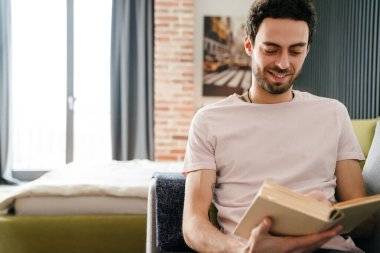 Mutlu traşsız adam gülümseyerek ve kitap okuyarak evde koltukta otururken