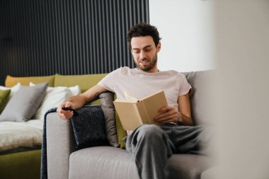 Evdeki kanepede kitap okurken uzaktan kumanda kullanan memnun bir adam.
