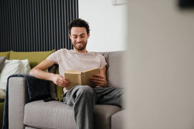 Mutlu traşsız adam gülümseyerek ve kitap okuyarak evde koltukta otururken