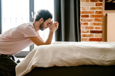 Evde, yatak odasında el ele tutuşup dua eden odaklanmış, traşsız bir adam.