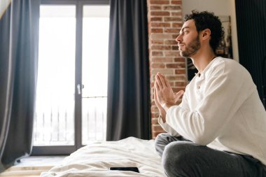 Odaklanmış esmer adam yatakta otururken meditasyon yapıyor ve cep telefonu kullanıyor.