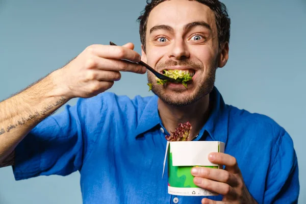 Excited handsome hungry guy eating salad takeaway isolated over blue background