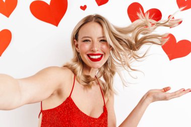 Heyecanlı güzel bir kız saçlarıyla dalga geçiyor ve beyaz arka planda izole edilmiş, kalpleriyle selfie çekiyor.