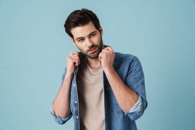 Mavi arka planda kameraya poz veren genç, yakışıklı, tıraşsız bir adam.