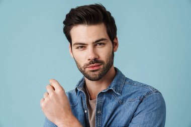 Mavi arka planda kameraya poz veren genç, yakışıklı, tıraşsız bir adam.