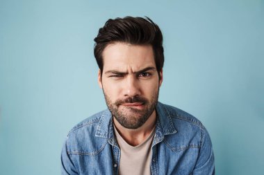 Genç, kafası karışmış, mavi arka planda yalnız bir şekilde poz veren ve kameraya bakan yakışıklı adam.