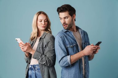 Genç ve şaşkın bir adam ve kadın mavi arka plandan izole edilmiş akıllı telefonlarını dikizliyorlar.