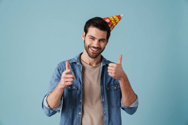 Genç yakışıklı, heyecanlı bir adam, parti konisinde, mavi arka planda izole bir şekilde baş parmak gösteriyor.