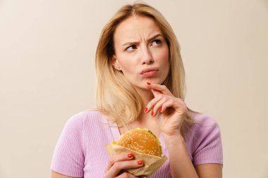 Düşüncelere dalmış güzel sarışın kız beyaz arka planda izole edilmiş hamburgerle poz veriyor.