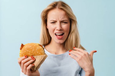 Mutlu olmayan güzel kız, mavi arka planda izole edilmiş hamburgerle poz verirken yüzünü buruşturuyor