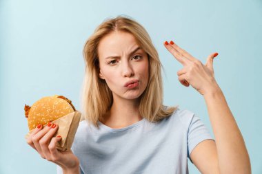 Mavi arka planda izole edilmiş hamburgerle poz veren mutsuz bir kız.