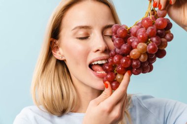 Mavi arka planda izole edilmiş üzüm yiyen mutlu sarışın kız.